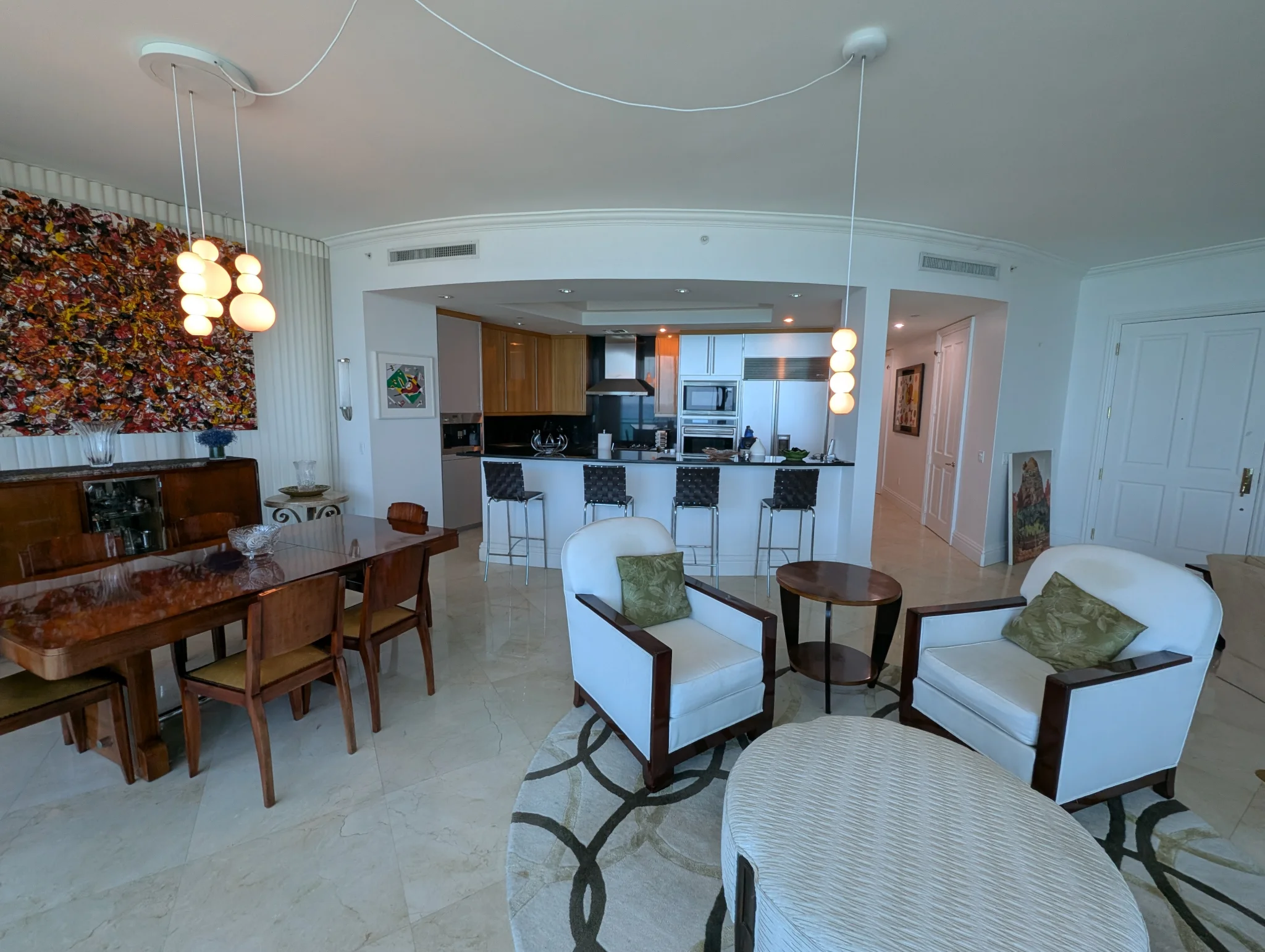 A wide shot of a modern, open-plan living and dining area with a kitchen island. The room features a large wooden dining table with chairs, a sleek white sofa, two accent chairs, and a kitchen with stainless steel appliances. A large, abstract painting hangs on the wall, and decorative pendant lights illuminate the space. The floor is covered in marble tiles, and a large circular rug with a geometric pattern sits in the seating area.