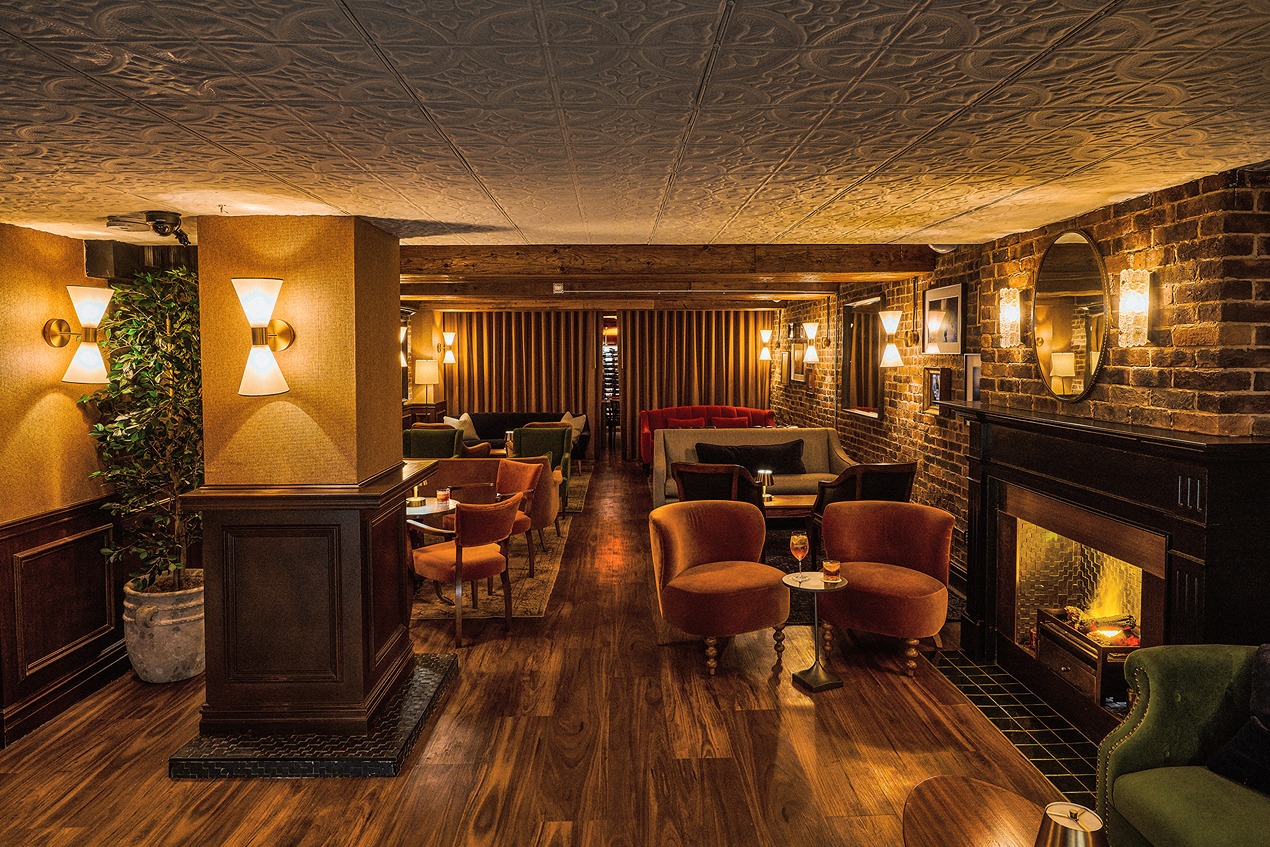 An elegant subterranean lounge featuring a brick fireplace, velvet armchairs in orange and green, a decorative tin ceiling, and warm amber lighting throughout the wood-floored space.