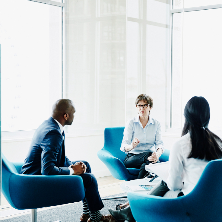 A group of four professionals sits in blue chairs in a modern office with large windows. They are engaged in a serious discussion, fostering a collaborative atmosphere.