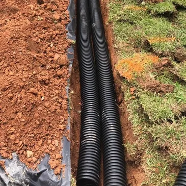 Technician installing a corrugated drainage pipe alongside a building foundation. Excavated soil, landscaping, and foundation waterproofing components are visible during the drainage system installation process.