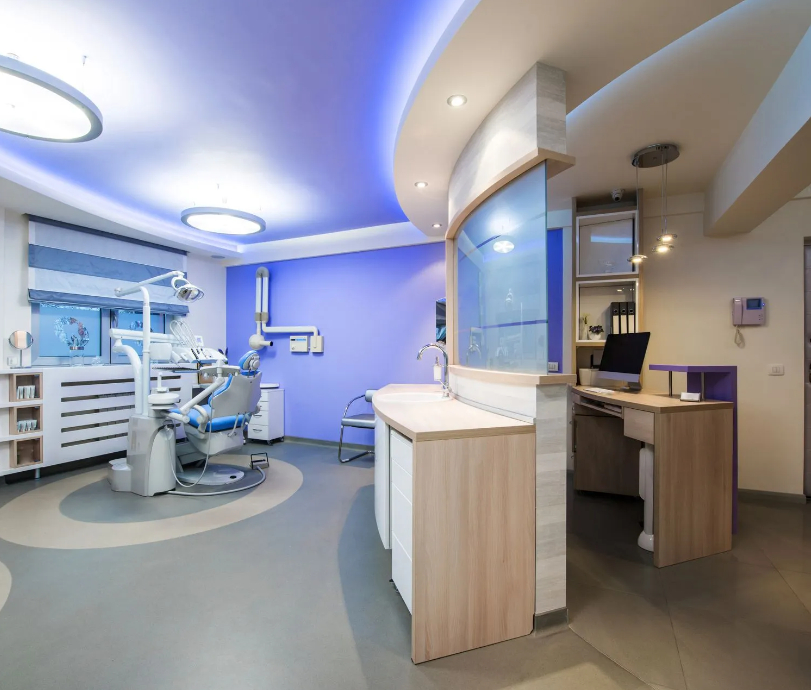 A modern, brightly lit dental operating suite featuring purple accent walls, a blue patient treatment chair, curved wooden cabinetry, ambient drop lighting, and a reception counter area.