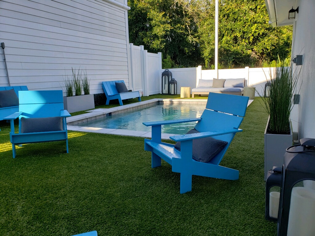 Small backyard pool area with artificial turf, bright blue Adirondack chairs, and a private residential setting.