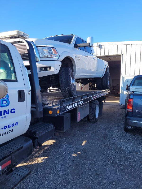 A low angle photograph captures a white lifted dually pickup truck mounted on a commercial flatbed tow truck outside an open auto repair shop under a vibrant clear blue sky.