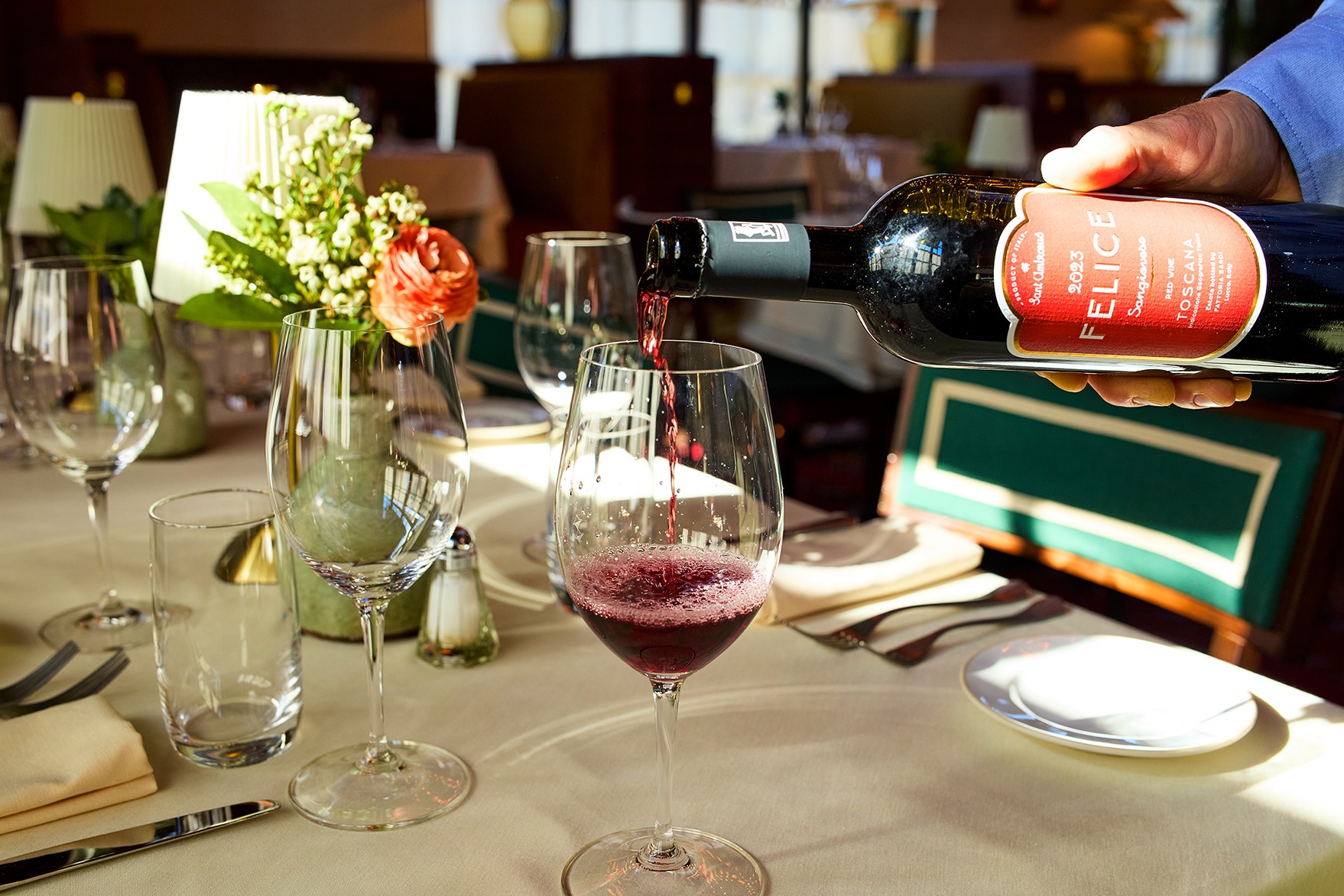 Red wine flows into a clear glass on a sunlit table. In the background, a small floral arrangement and a pleated lamp create an elegant dining atmosphere.