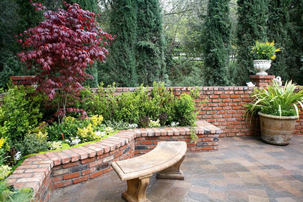 Serene brick garden patio with curved bench, lush flower beds, red-leaf tree, potted plants, and tall evergreens in the background.