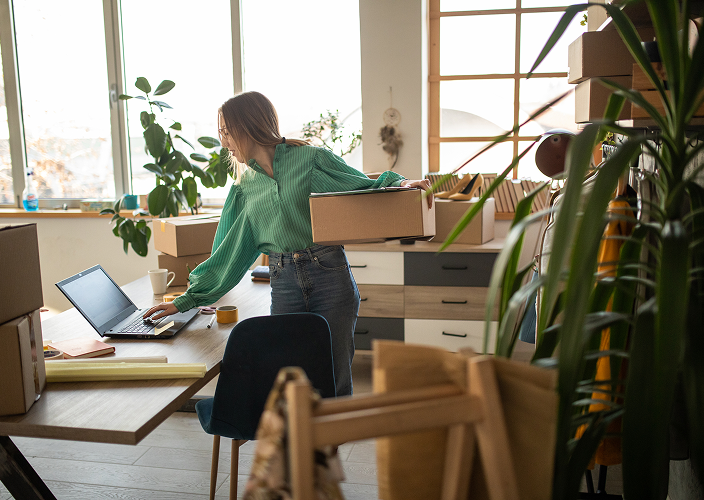 A woman multitasking in her new office;  working on her computer with one hand and holding a box in the other.