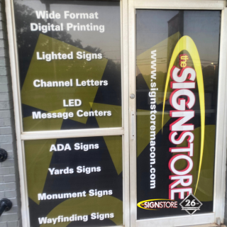 Window display of Signstore listing services: Wide Format Digital Printing, Lighted Signs, Channel Letters, LED Message Centers, ADA Signs, Yards Signs, Monument Signs, and Wayfinding Signs. Signstore logo and "26" anniversary badge are visible.