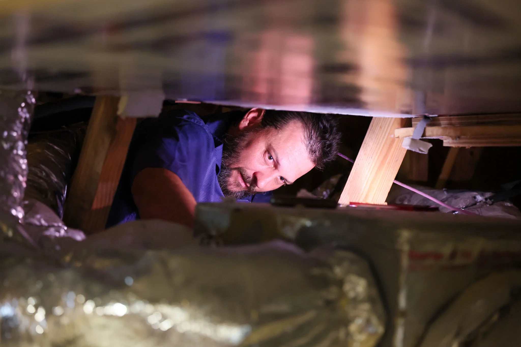 A man with a beard wearing a blue shirt is looking down into a tight space between wooden beams and  