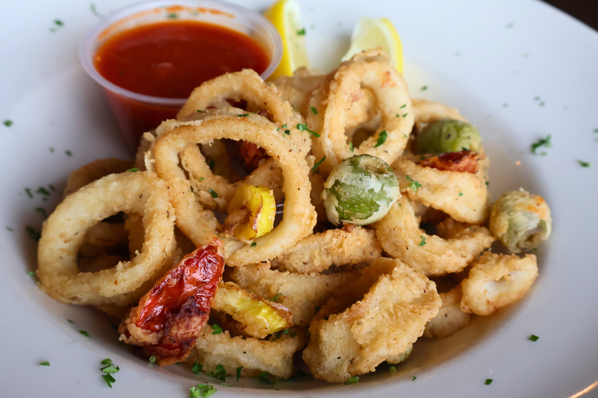 Golden fried calamari paired with fried green olives, roasted tomatoes, and a side of marinara sauce for dipping, offering a savory and tangy appetizer.