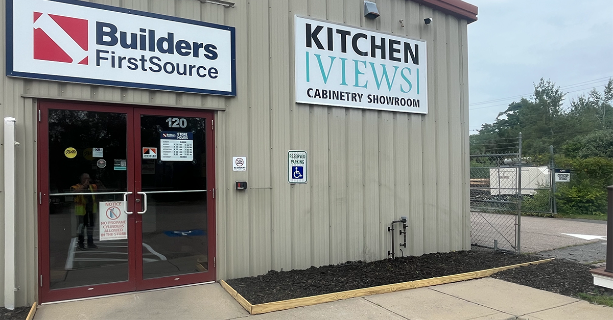 Exterior entrance of a Builders FirstSource store and Kitchen Views cabinetry showroom, showing red double doors with posted store hours, an accessible parking sign, and a landscaped mulch bed along the building front.