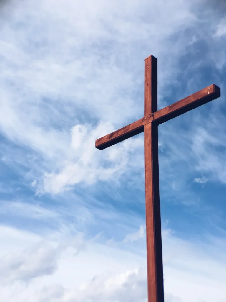 A dark brown wooden cross stands against a bright blue sky with wispy white clouds.