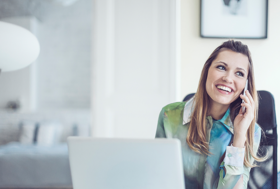 A woman in a colorful blouse smiles while talking on her phone at a desk. A blurred background with a framed picture adds to the casual atmosphere.