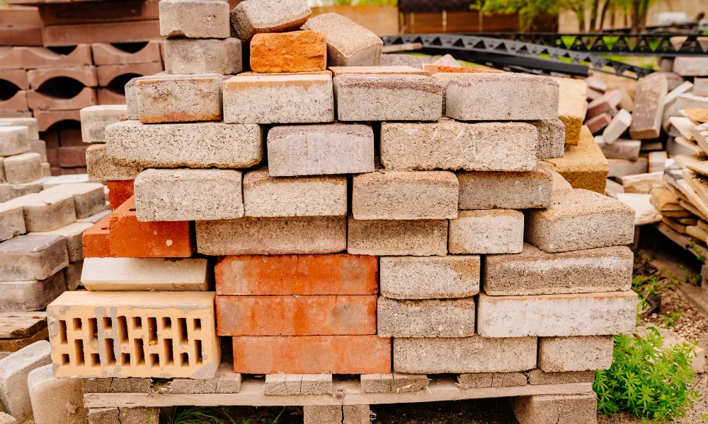 A close-up view of stacks of bricks and paving stones on a wooden pallet. The bricks are of various sizes, shapes, and colors, including red, grey, and beige. Some bricks are solid, while others have holes. In the background, more stacks of building materials are visible, along with some greenery and a wooden fence.