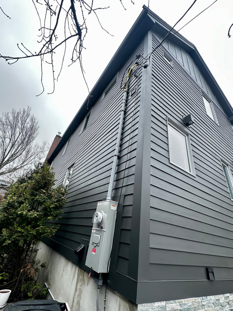 A modern, dark gray house with horizontal siding and various electrical conduits and a meter box attached to the exterior wall. The house has several windows and a prominent corner. Bare tree branches extend into the frame from the upper left, and a green conifer tree is visible on the left side of the house. The sky is overcast.