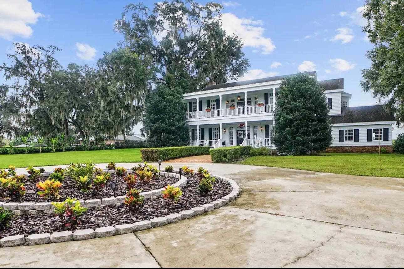 Exterior view of a custom-built luxury farmhouse in Central Florida featuring a wraparound porch, landscaped entrance, and expansive property, showcasing the craftsmanship of custom home builders in Central Florida.