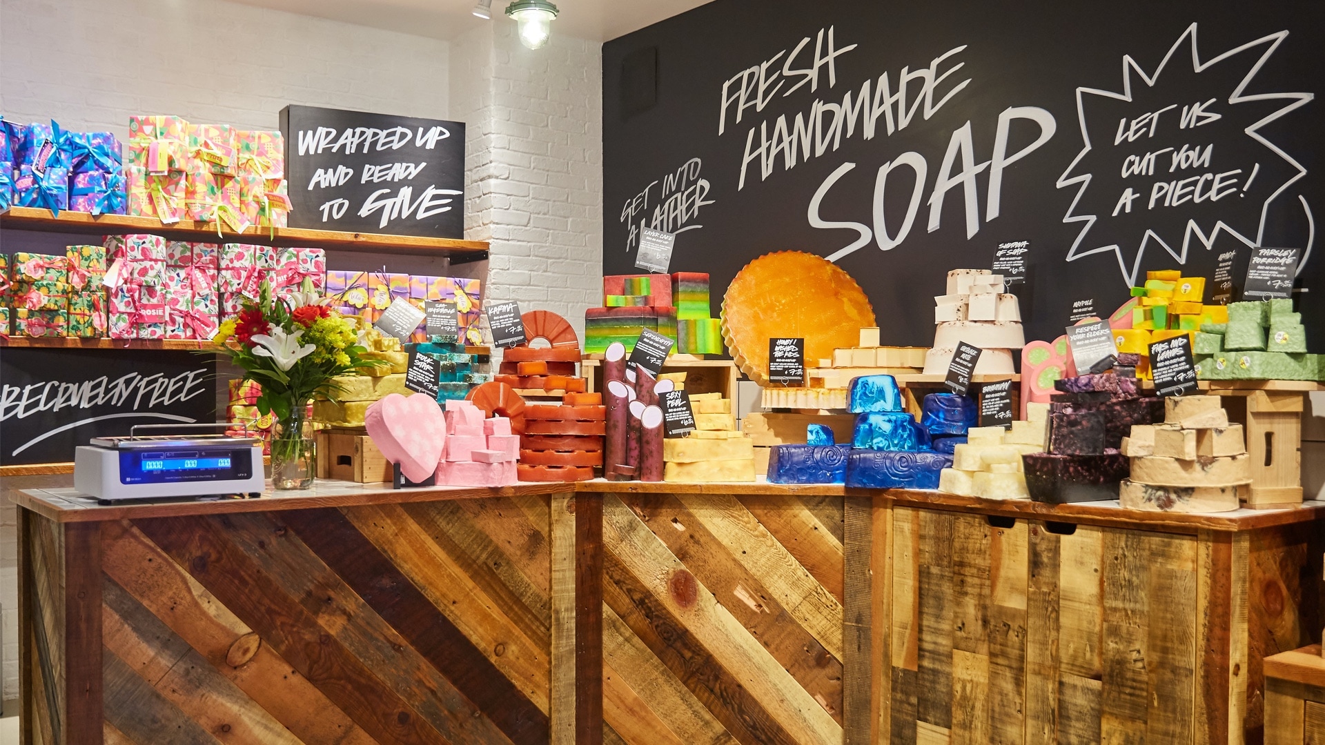 An einer Ecke eines LUSH-Ladens steht ein großes Schild auf dem geschrieben steht: „Fresh Handmade Soap“ und in kleineren Buchstaben „Let us cut you a piece“. Auf einem Tisch liegen verschiedene Farben und Größen von handgemachten LUSH-Seifen.