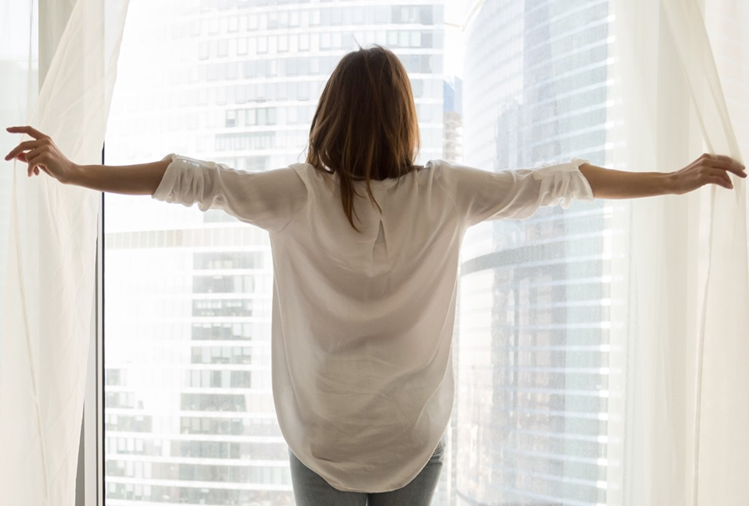 Woman opening white curtains, standing with arms outstretched, facing tall city buildings. The scene conveys a sense of freedom and morning light.