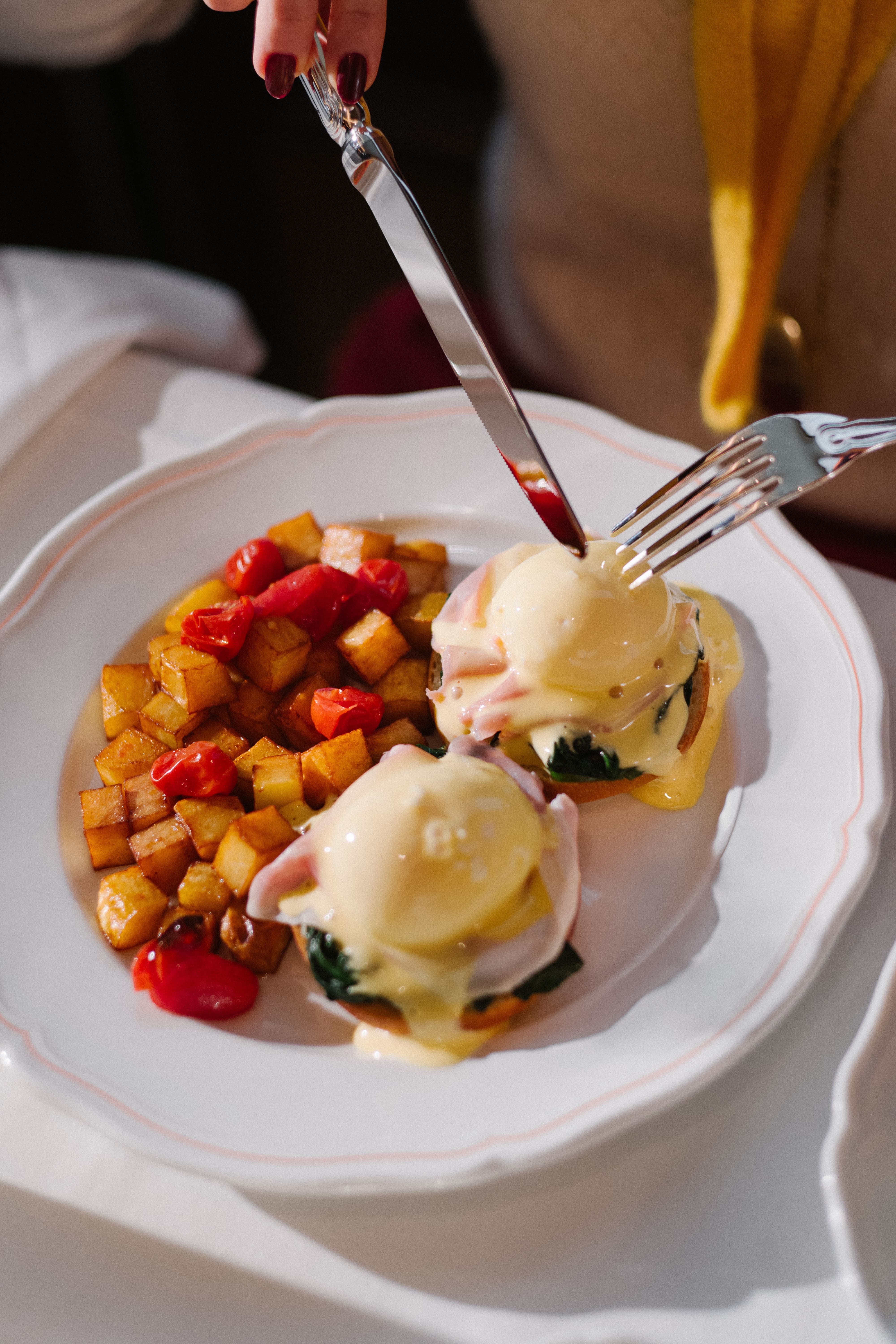 Close-up of eggs Benedict on a white scalloped plate—two poached eggs with hollandaise sauce over greens and ham, served with golden diced potatoes and cherry tomatoes, as a hand cuts into one egg with a knife and fork.