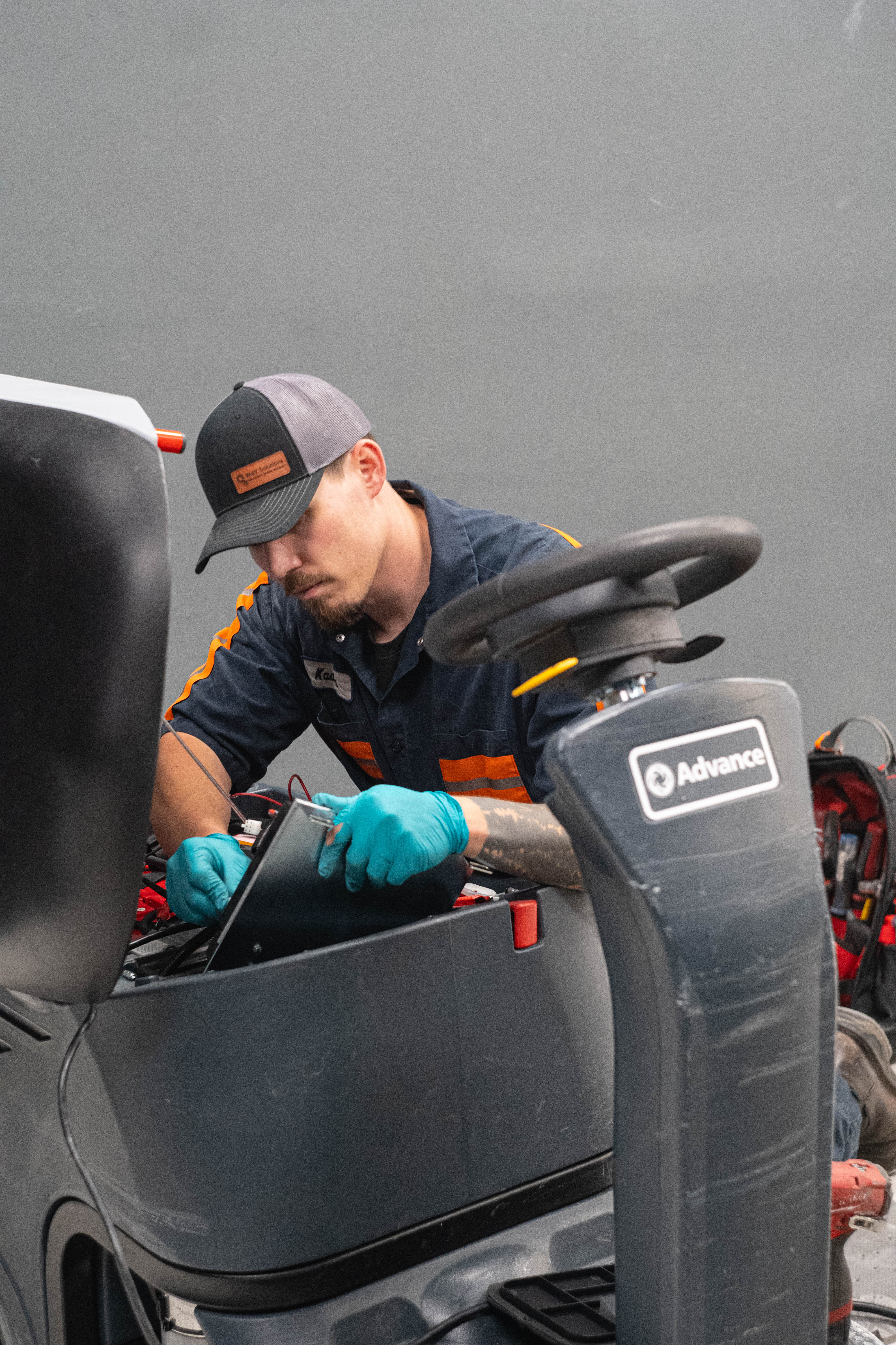 A WAY Solutions technician wearing branded gear performing complex electrical wiring diagnostics and battery testing on a heavy-duty Advance ride-on floor scrubber.