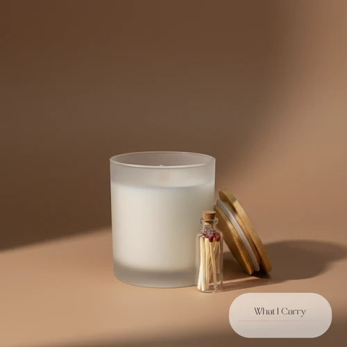 A frosted glass candle with a wooden lid, a small glass bottle filled with matches, and the text "What I Carry" are arranged on a plain, neutral-colored surface. The lighting creates a soft shadow on the left side of the image.