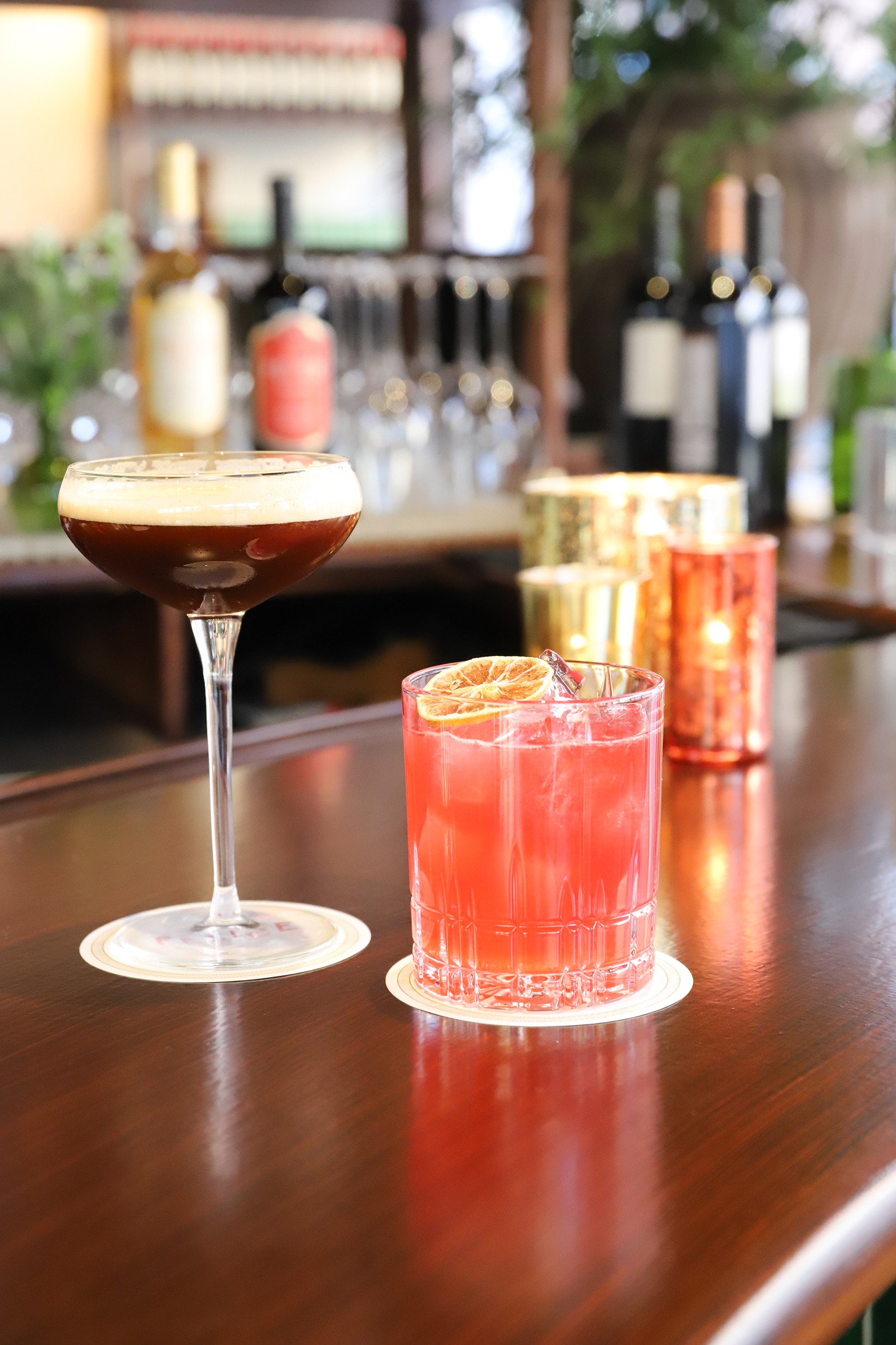An espresso martini with a thick foam head and a vibrant red cocktail garnished with a dried orange slice. The background shows an upscale bar with various wine bottles.