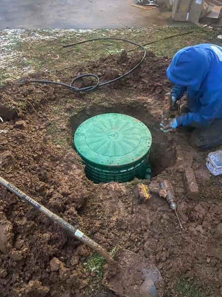A worker in a blue hoodie kneels in mud to install or repair a green septic tank riser. Tools, including a shovel and power drills, surround the excavation site.