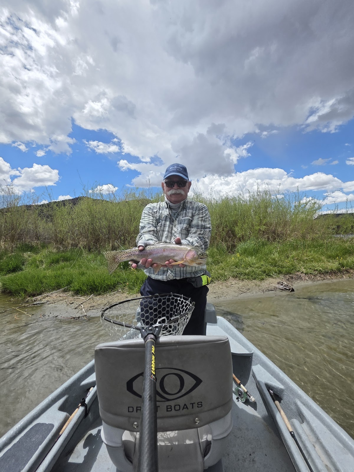 A man in a plaid shirt and baseball cap holds a rainbow trout he caught while standing in a boat on a river. The boat has a logo that reads "DR T BOATS" and a fishing net. The background shows green brush and a cloudy blue sky.