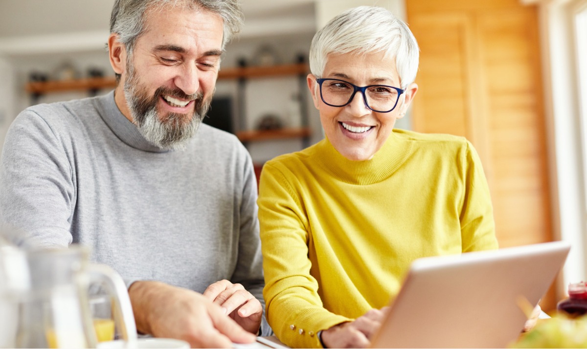 A middle-aged couple, a man with a grey beard and a woman with short white hair and glasses, are smiling while looking at a laptop. They are sitting at a table with a pitcher of orange juice and a jar of jam. The woman is wearing a yellow turtleneck sweater, and the man is wearing a grey turtleneck sweater. They appear to be in a home environment.