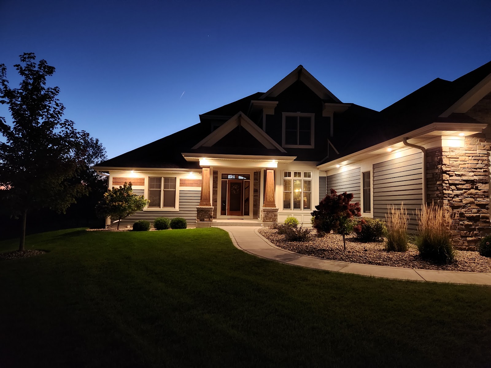 A well-lit, two-story house with blue siding and stone accents is illuminated at dusk. A pathway leads to the front door, surrounded by manicured landscaping and a lush green lawn. The sky is a deep twilight blue, with a faint airplane contrail visible.
