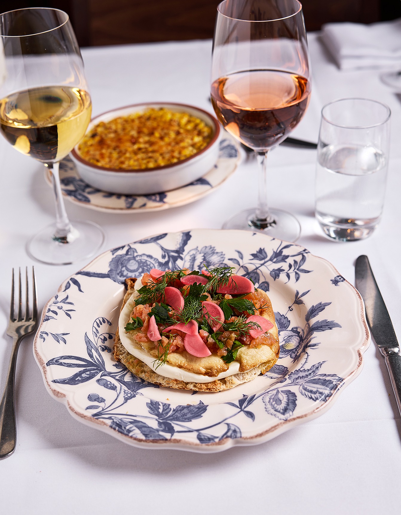 An elegant seafood bruschetta topped with pickled radishes and fresh dill, served on a blue floral patterned plate with wine pairings on a white tablecloth.