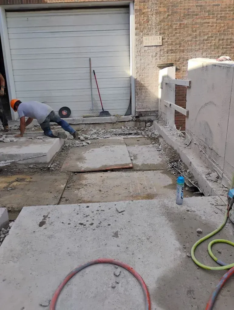 Worker removing broken concrete slabs near garage entrance, surrounded by debris, tools, and hoses,  