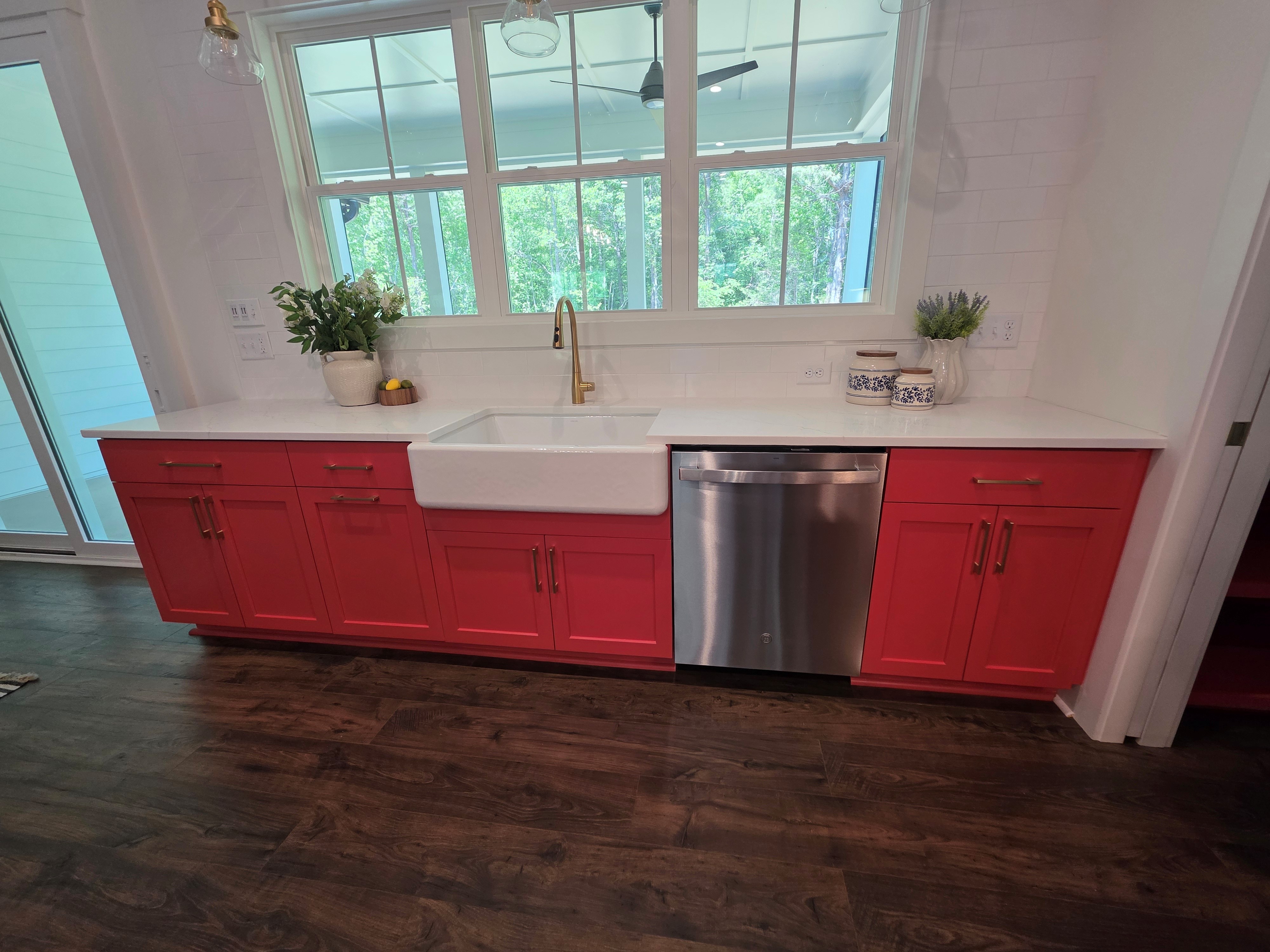 Bright kitchen with red base cabinets, white farmhouse sink, stainless steel dishwasher, white tile backsplash, and large windows overlooking trees.