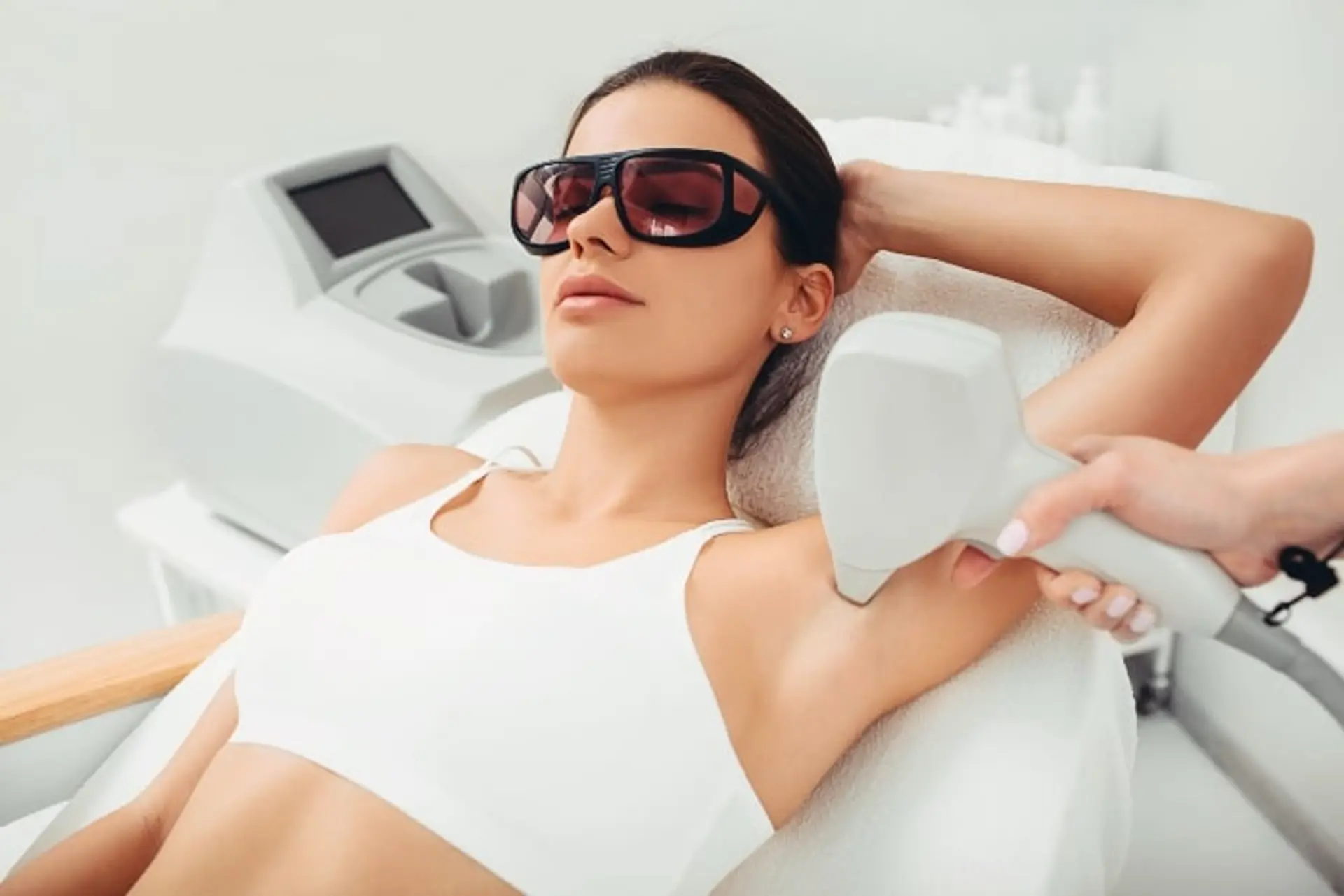 A woman wearing protective goggles lies down while undergoing laser hair removal on her armpit. A person's hand holds a laser device to her skin, with a medical machine in the background.