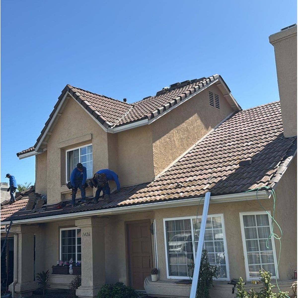 Angle view of professional roofers wearing blue shirts working on the brown concrete tile roof of a two-story beige stucco house under a bright sunny sky.