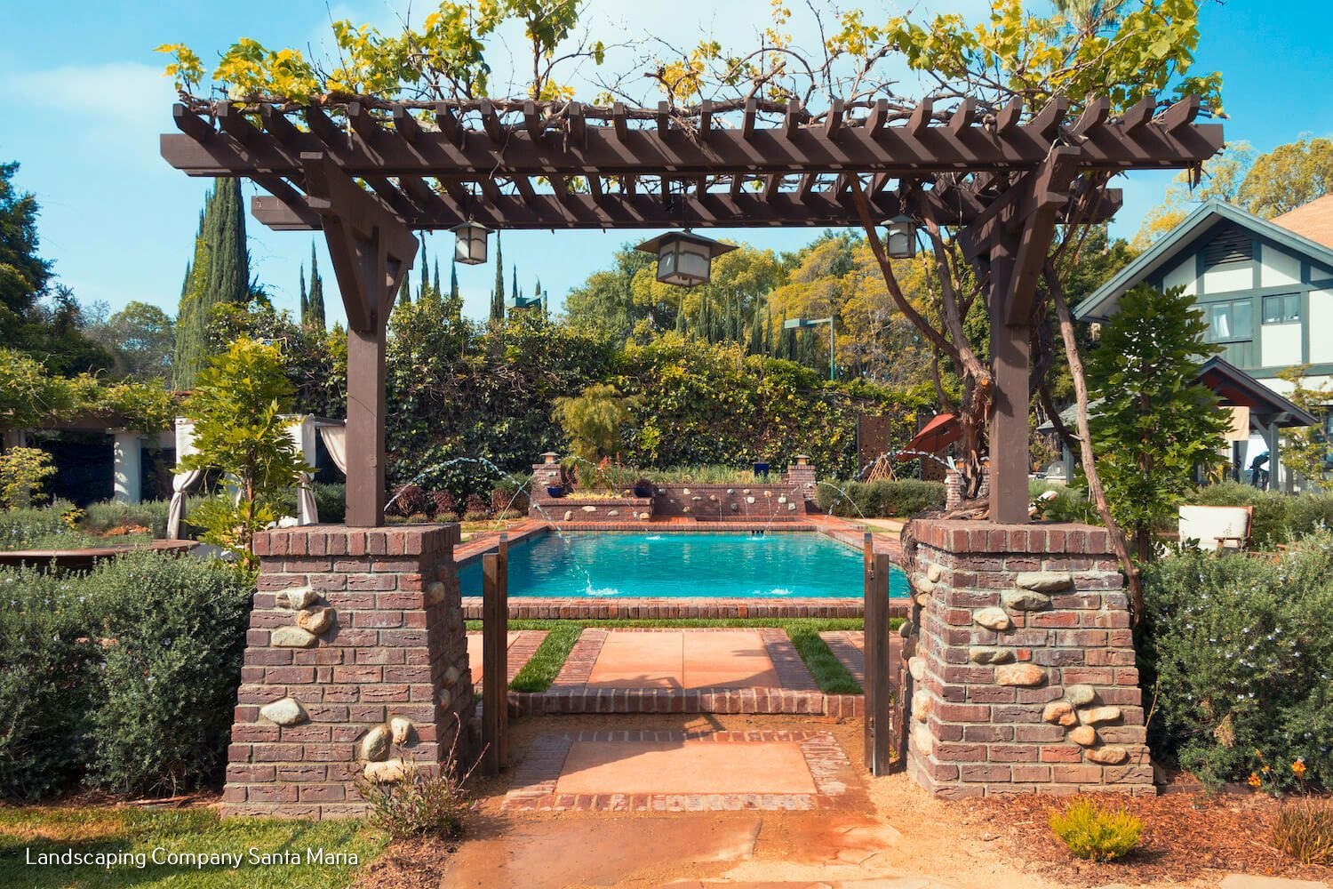 Rustic wooden pergola frames a bright turquoise pool, lush garden, brick path, and welcoming home in a sunny landscape.