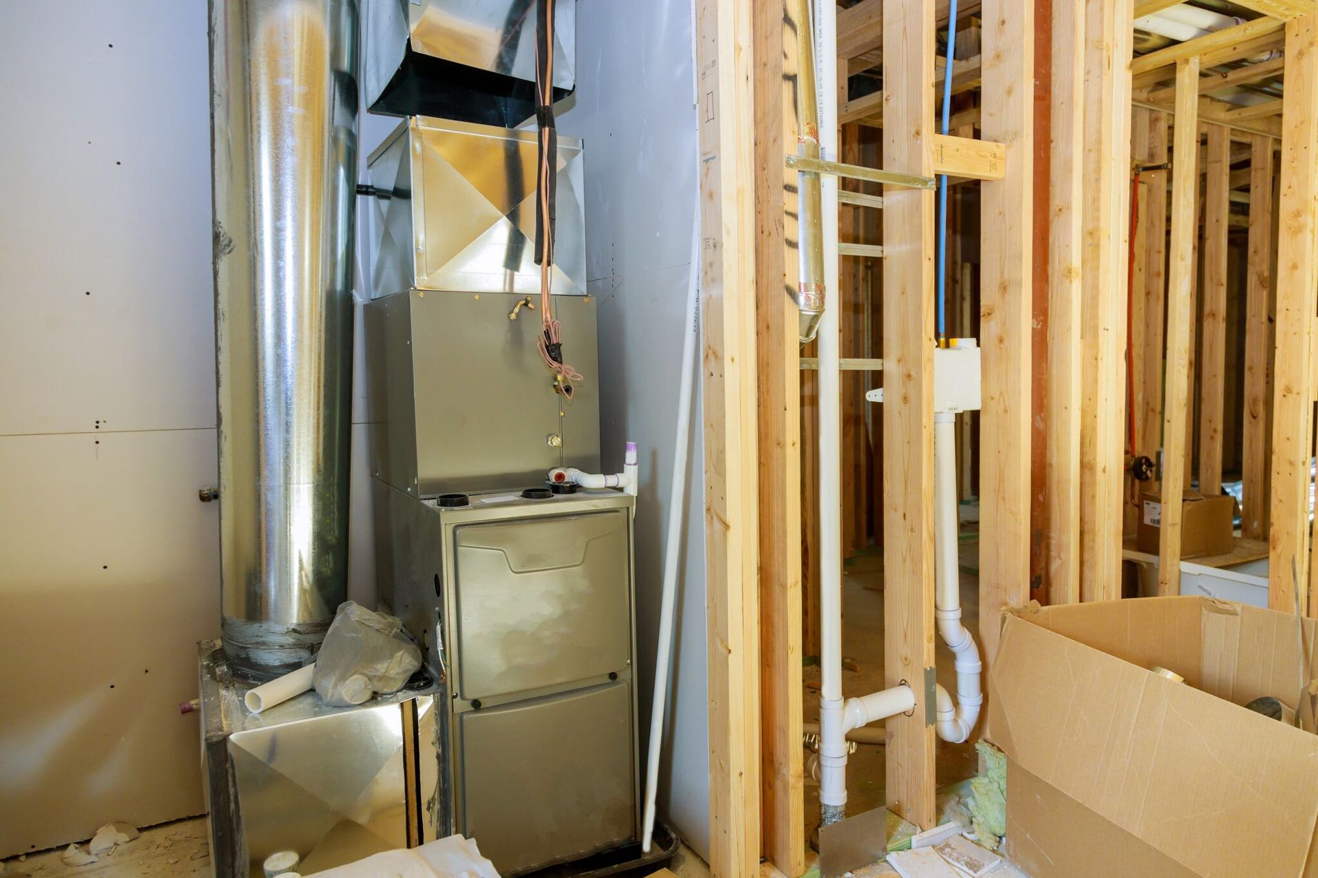 New HVAC furnace installed in unfinished utility area with exposed wooden framing, ductwork, and plumbing pipes, illustrating residential heating system setup during construction, including ventilation connections and mechanical components.
