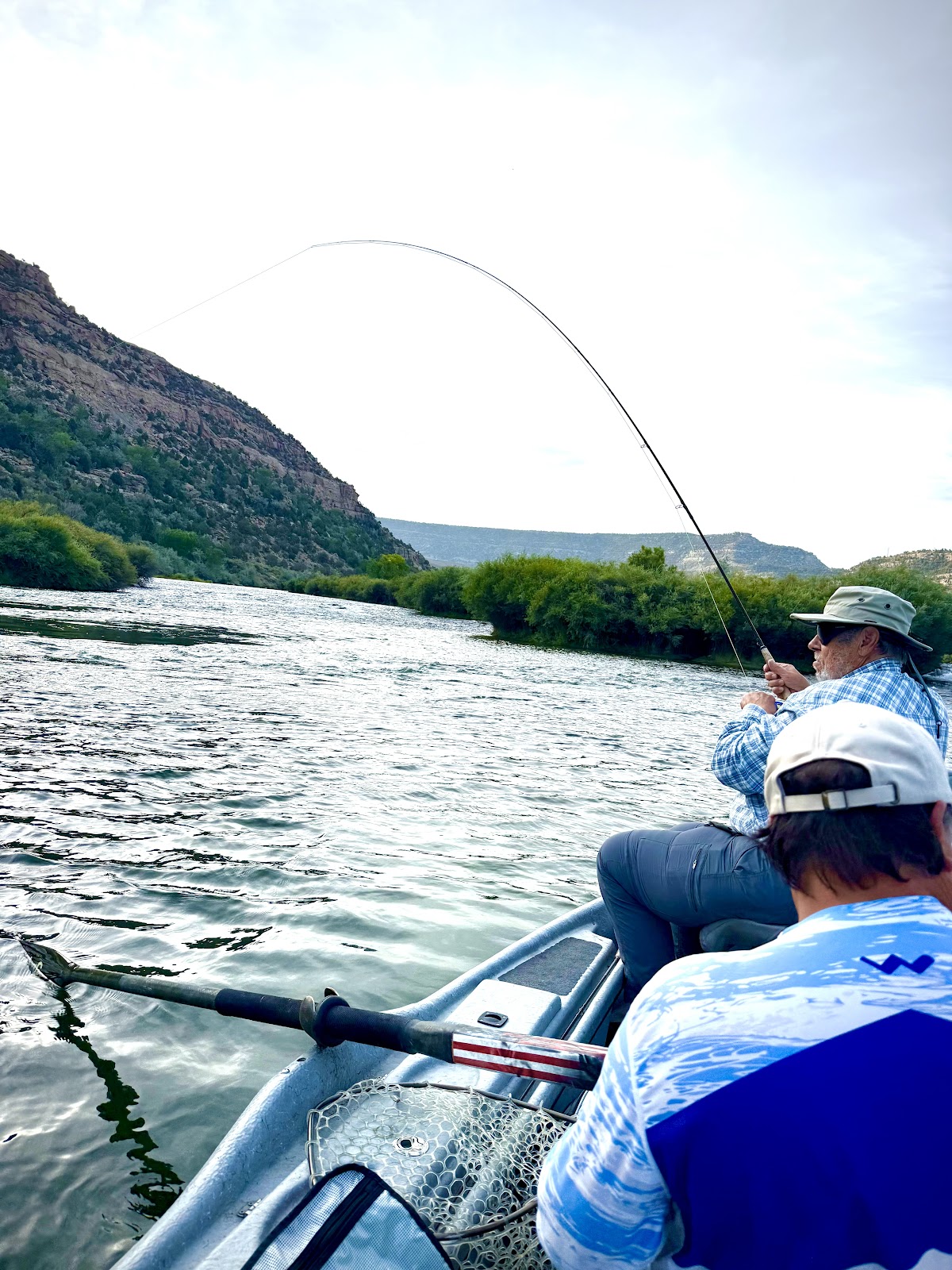 Two people are fishing from a boat on a river. The person in the foreground is wearing a white baseball cap and a blue and white patterned shirt. The person in the background is wearing a hat and sunglasses and a blue and white plaid shirt. They are both holding fishing rods, and the rod of the person in the background is bent, indicating they may have a fish on the line. The river is flowing and surrounded by green vegetation and rocky hills. The sky is overcast.