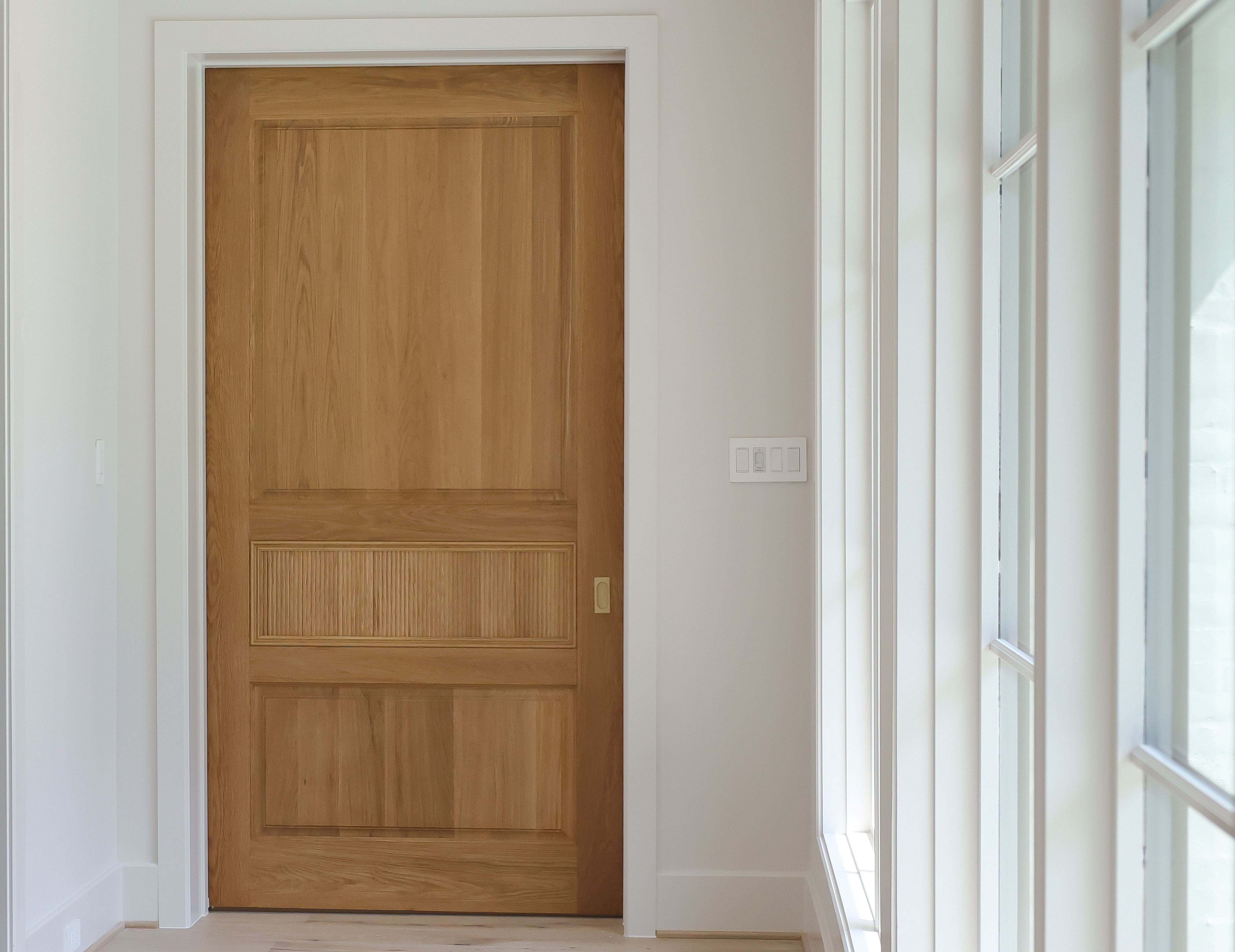 A sleek white oak pocket door with a natural wood finish, elegantly closed within a light-filled hallway.