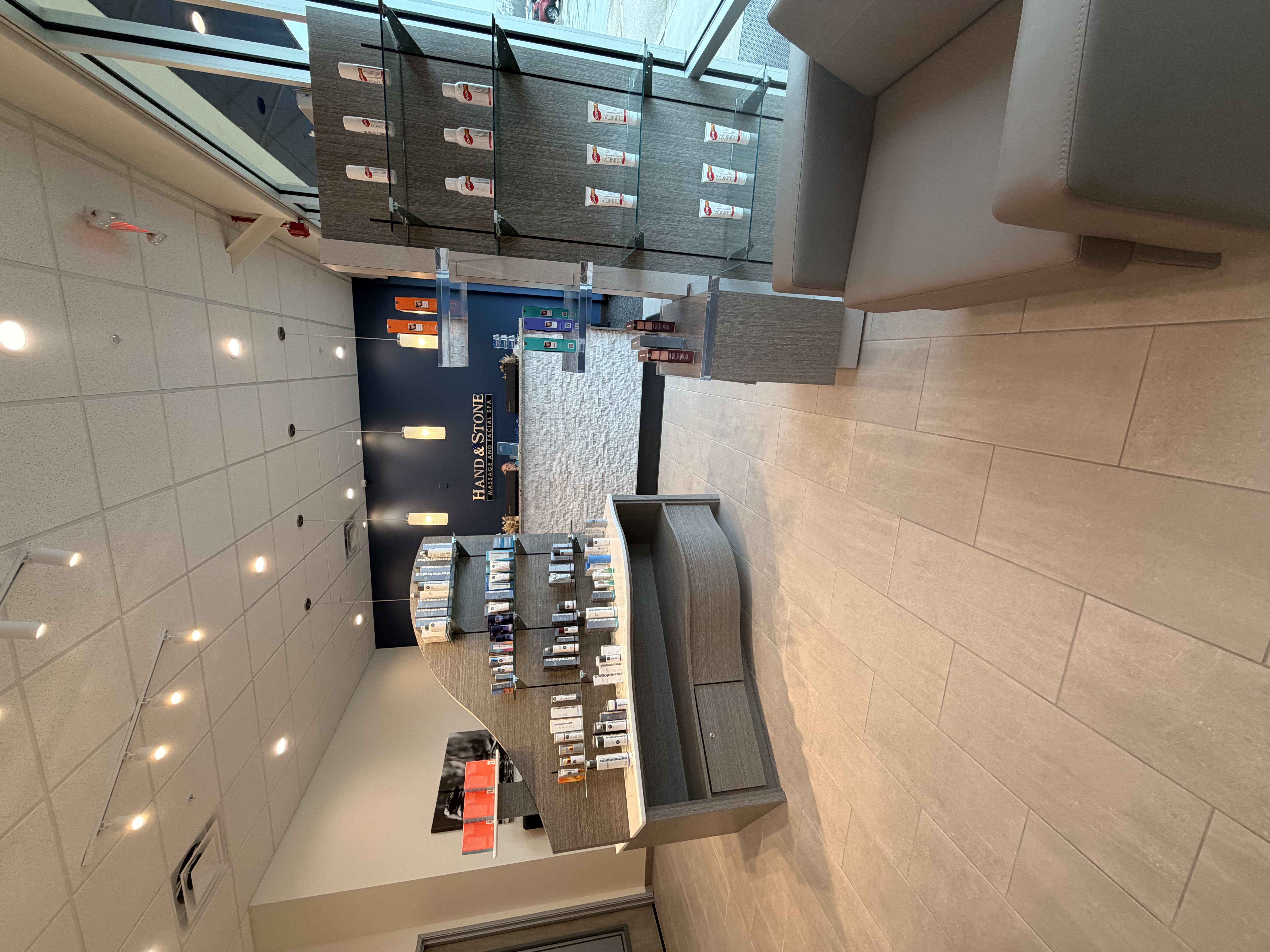 Elevated view of reception desk with glass shelving displaying skincare products. Gray wood-grain cabinetry with white countertops. Stacked stone accent wall and navy blue Hand & Stone logo wall. Track lighting and pendant fixtures illuminate space.