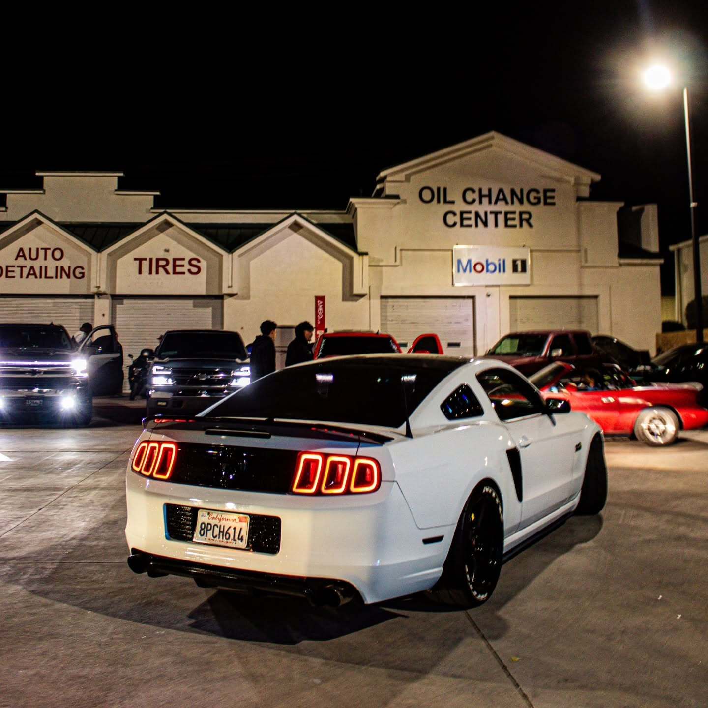 White Ford Mustang parked outside an illuminated oil change center during a nighttime automotive mee 