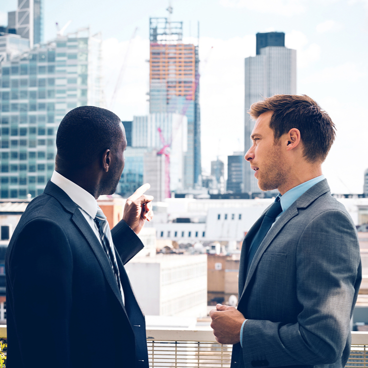Two men in suits stand on a rooftop, discussing while looking at a city skyline with modern skyscrapers. The scene conveys a professional tone.
