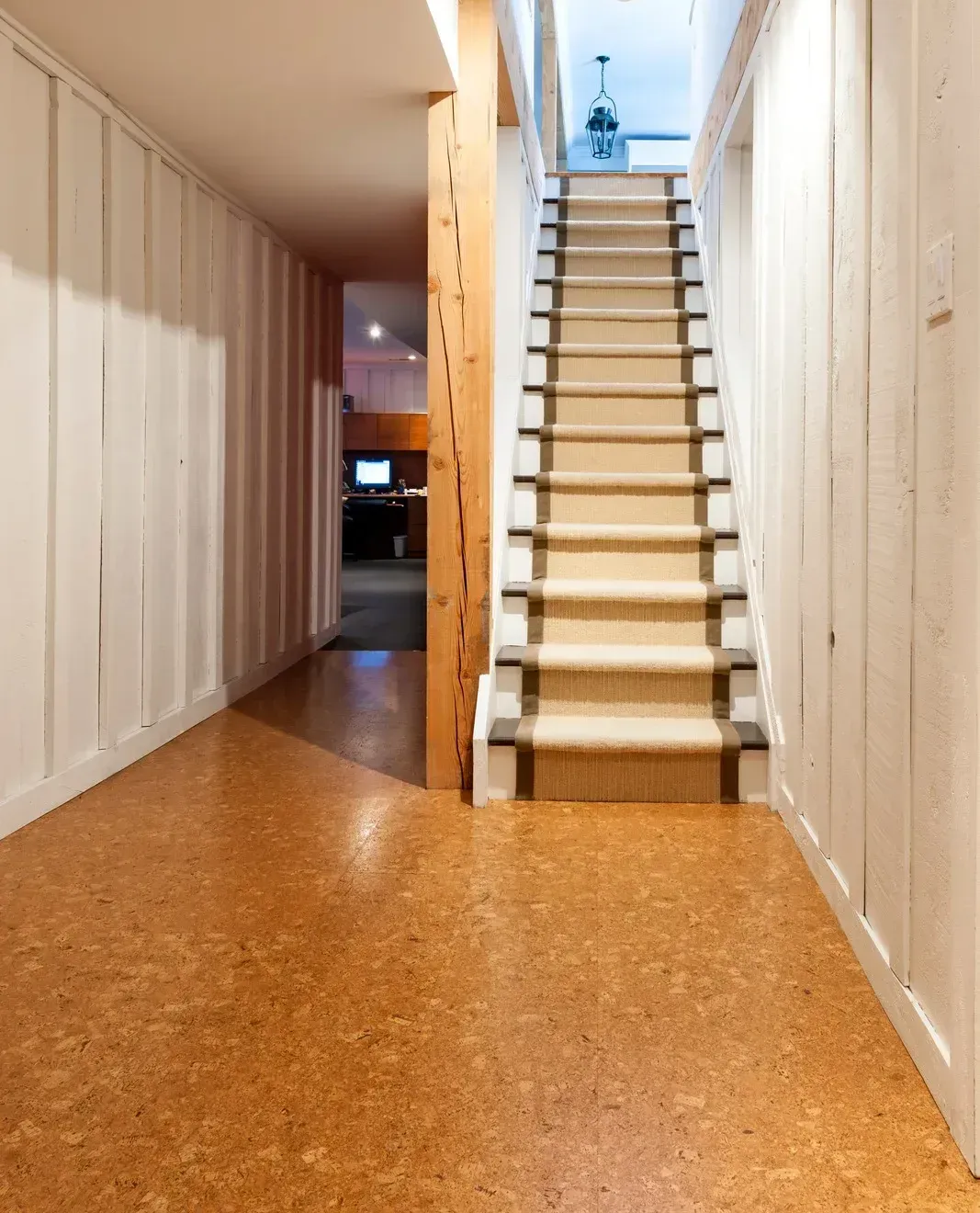 An interior view of a hallway with cork flooring leading to a carpeted staircase, with white paneled walls and a wooden post.