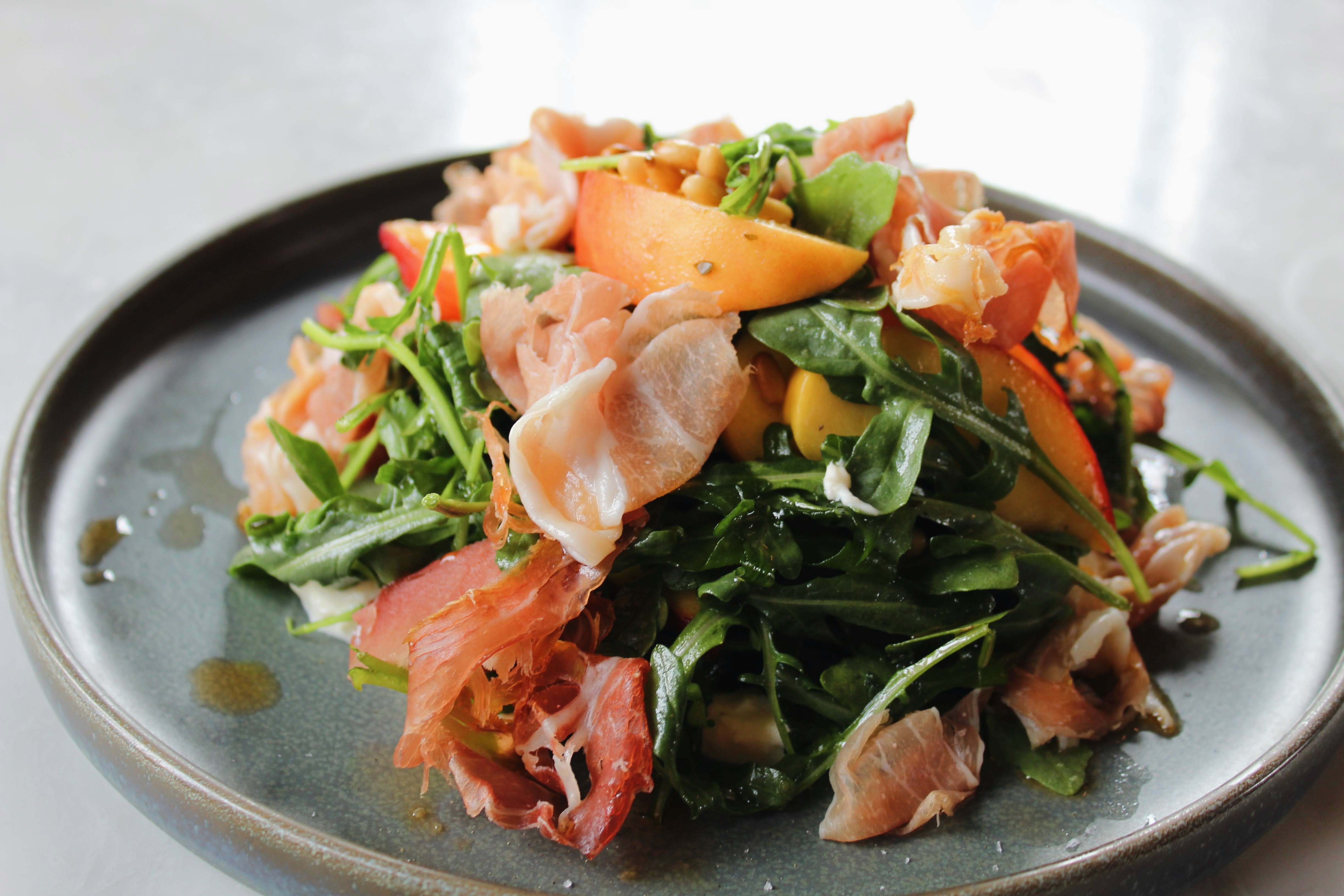 Peach & Prosciutto Salad — ripe summer peaches and ribbons of prosciutto over peppery arugula with stracciatella, mint, toasted pine nuts, white balsamic vinaigrette, and Calabrian chili oil.