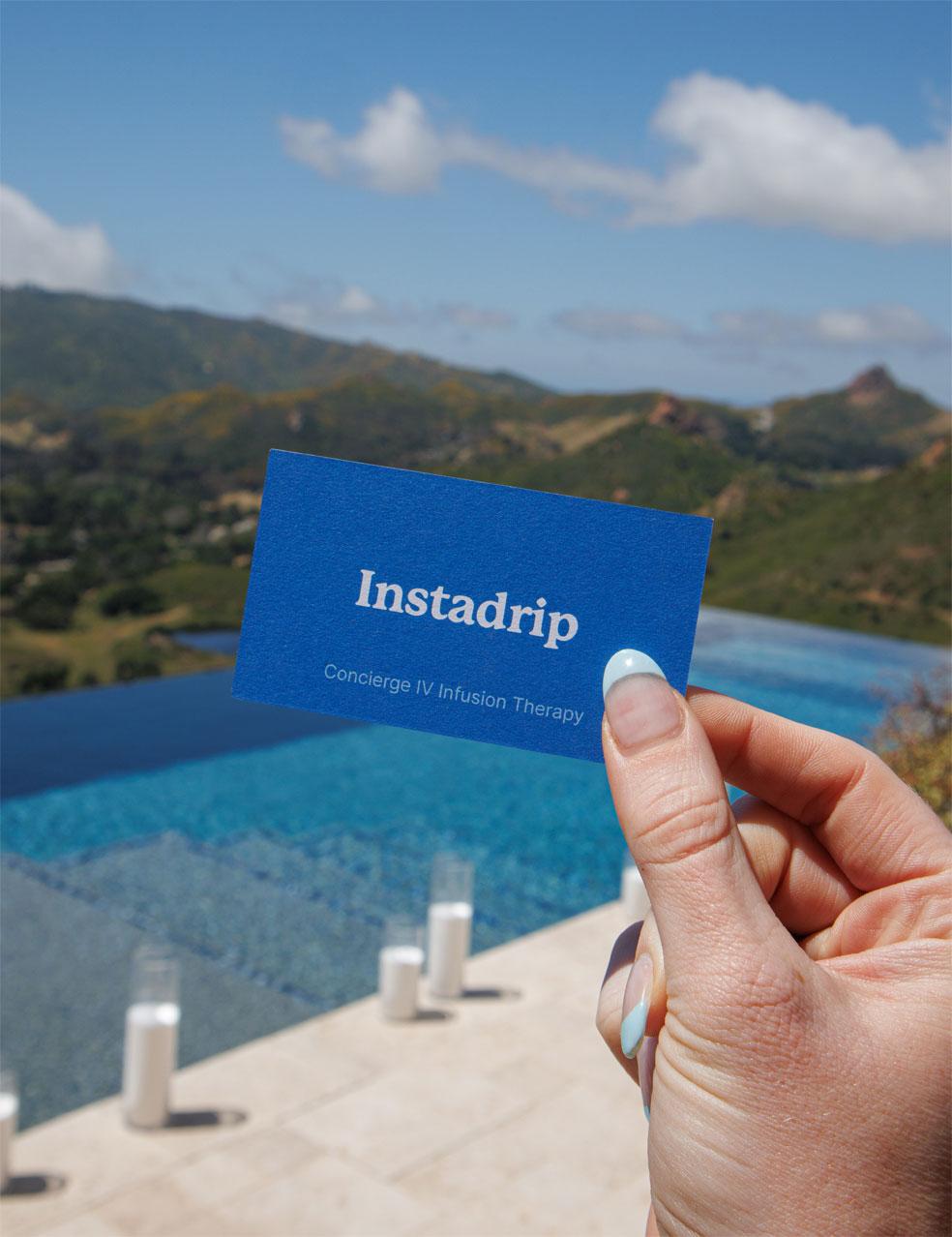 The photo shows a hand holding an Instadrip business card with the words “Concierge IV Infusion Therapy,” in front of a bright blue pool. The background includes green hills and cylindrical white candles by the water’s edge.