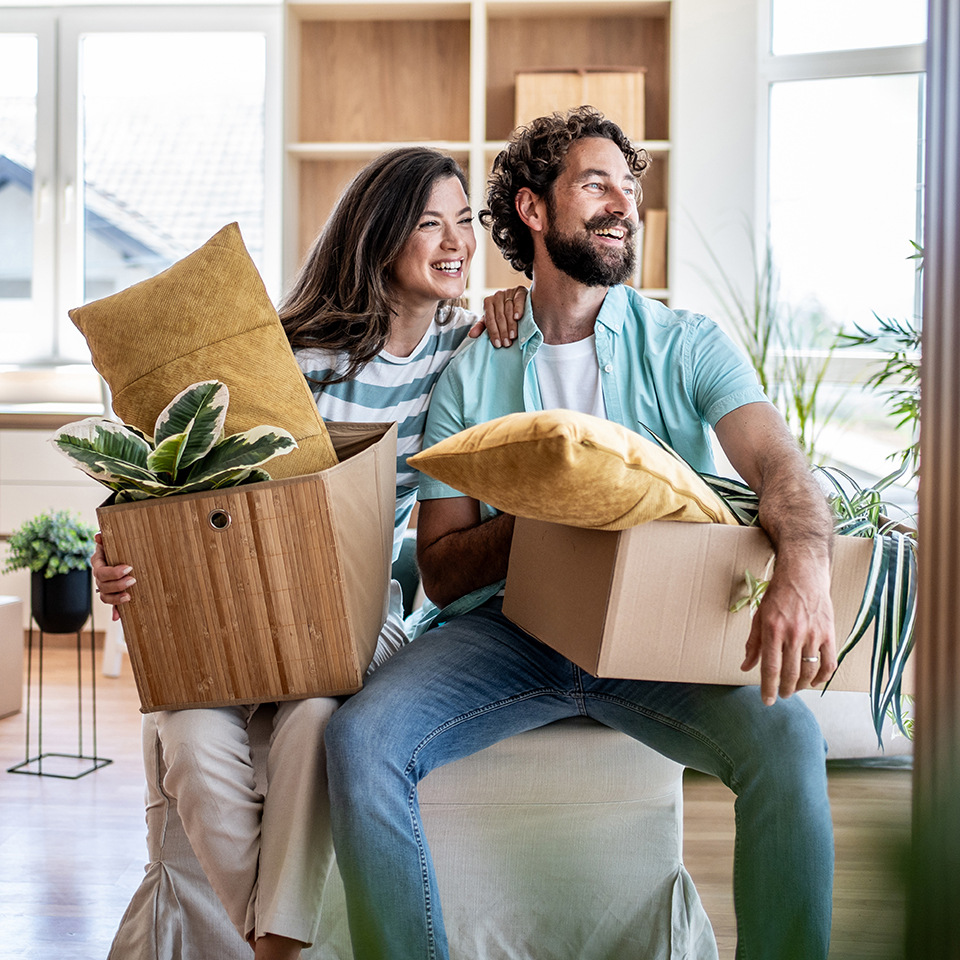 A woman and man sitting on an ottoman holding boxes containing pillows and plants.