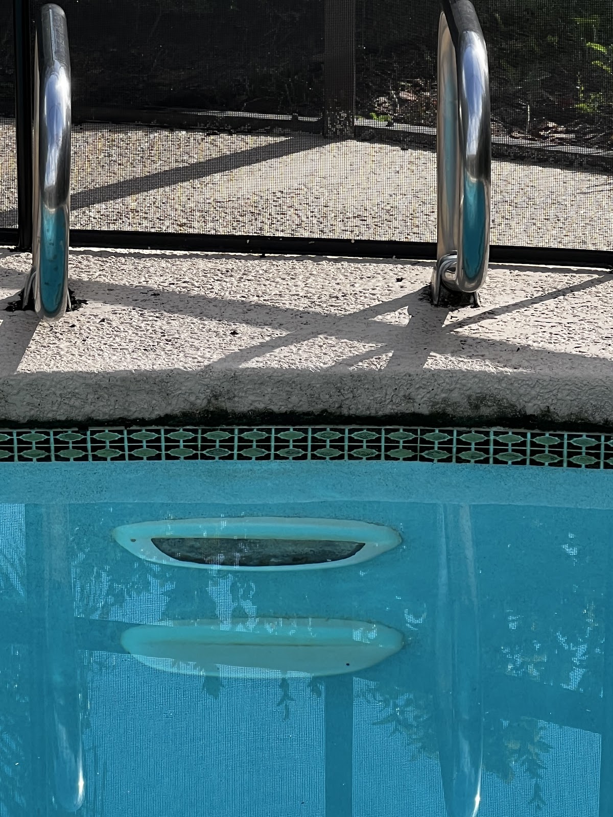 Close-up of a swimming pool skimmer opening beneath a stainless steel ladder, with clear blue water and decorative tile.