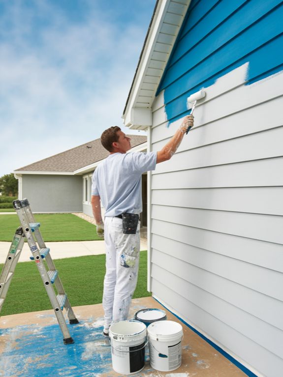 A painter in white work clothes is painting the side of a light gray house with blue paint using a r 