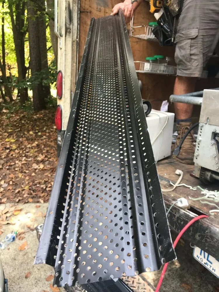 A worker holds a black perforated gutter guard while standing on a work truck, surrounded by tools and equipment in a wooded area.