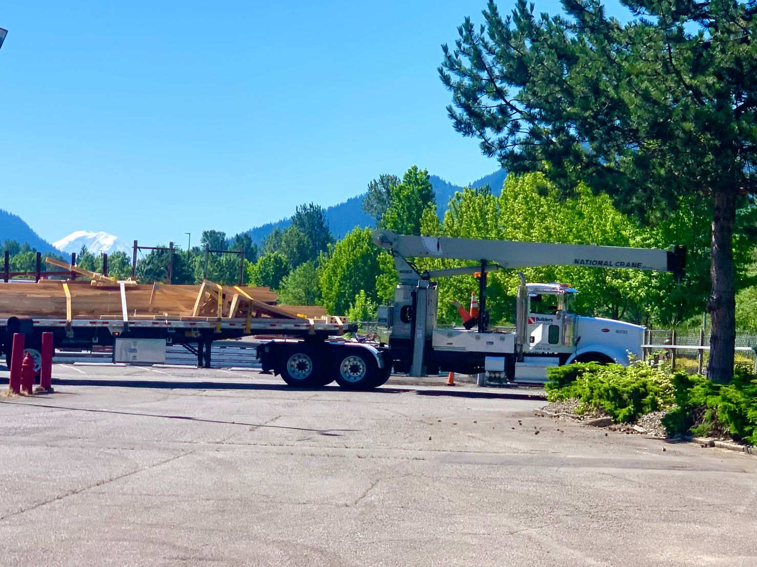 A Builders FirstSource delivery truck with a crane, loaded with lumber, parked in the yard. The scene includes vibrant green trees and a distant view of a mountain under a bright blue sky.