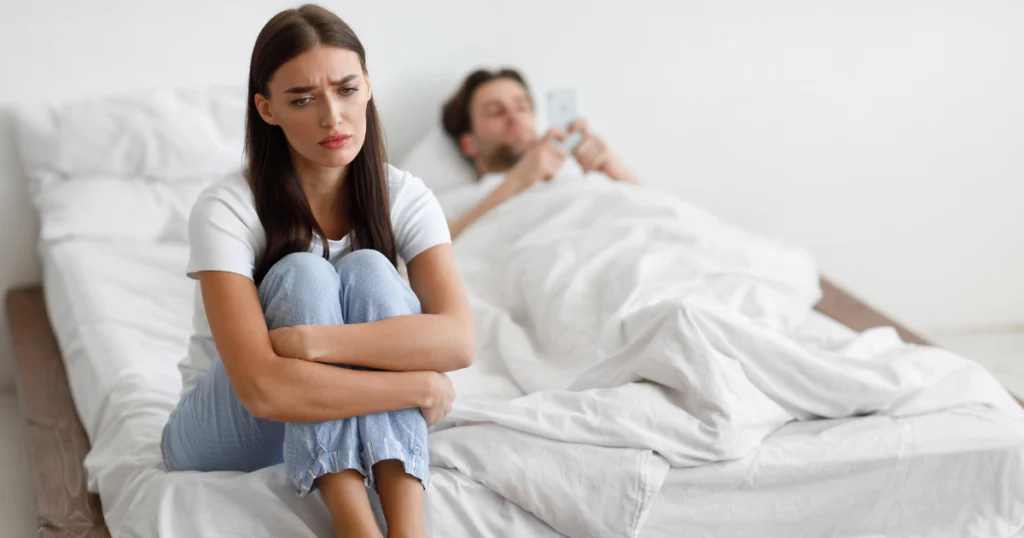 A distressed woman sits on the edge of a bed, hugging her knees, while a man lies in the background, engrossed in his phone. The setting appears to be a bedroom, with white bedding and walls. The woman's body language and facial expression convey sadness and isolation, while the man's preoccupation with his phone suggests a disconnect between the couple.